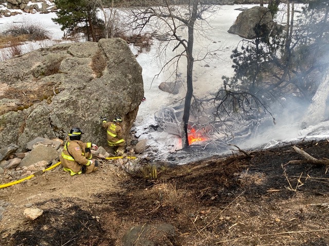 Firefighters are extinguishing a small wildfire near a frozen lake, amid rocks and trees.