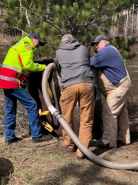 Three individuals working with a large hose next to a metal structure outdoors.
