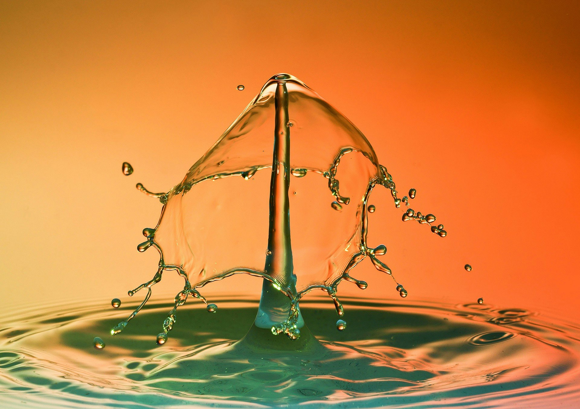 A high-speed photograph captures a water drop collision, creating a unique liquid sculpture effect against an orange backdrop.