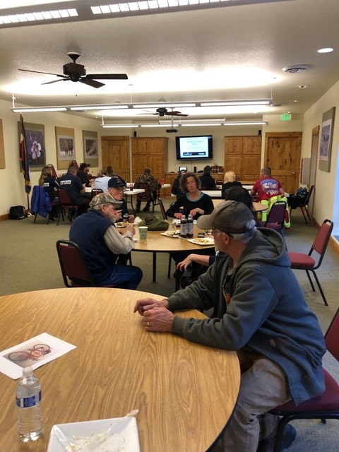 A group of people seated at tables in a room with images on walls and a screen displaying information.
