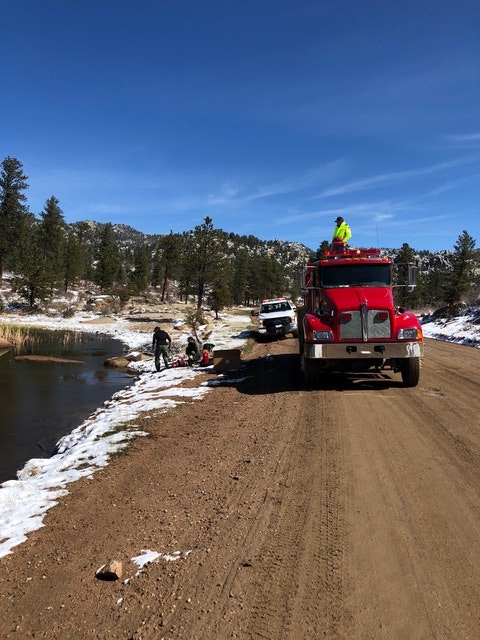 A fire truck on a dirt road with people working near a water body, patches of snow, and a clear blue sky.