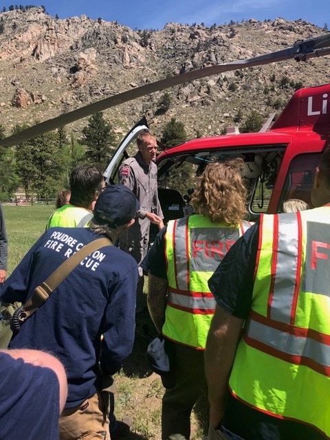 Group of people with "FIRE RESCUE" vests listening to a person by a red helicopter, amidst a natural, rocky landscape.