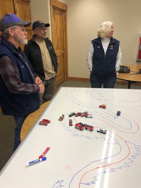 Three people observing toy cars on a table with drawn tracks, possibly for a planning or demonstration purpose.