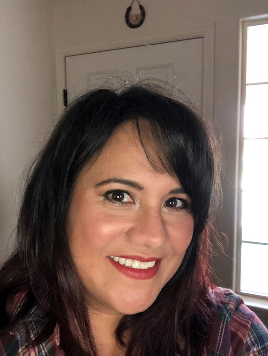 A person smiling indoors, with dark hair and wearing a plaid shirt.