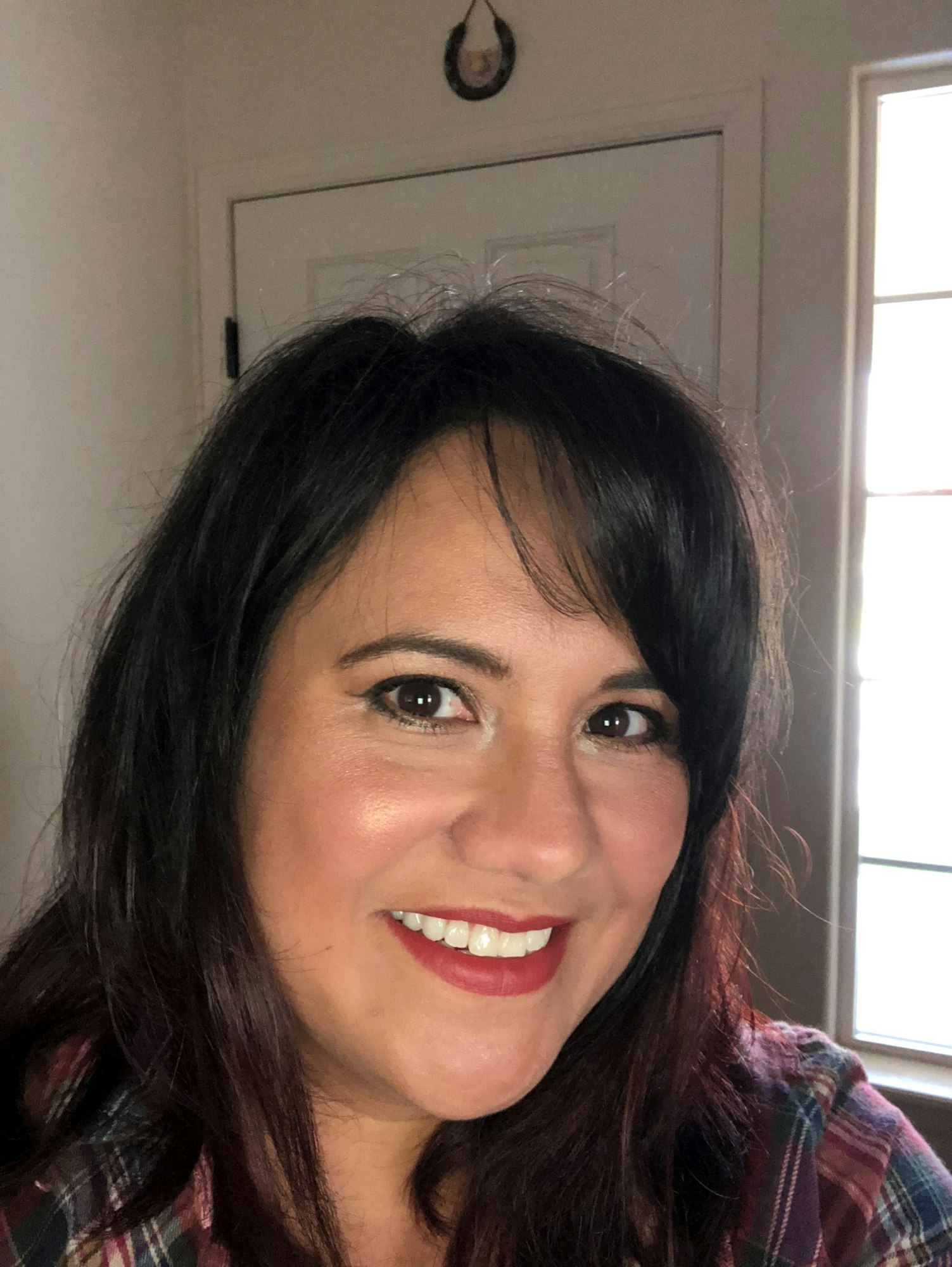 A person smiling indoors, with dark hair and wearing a plaid shirt.
