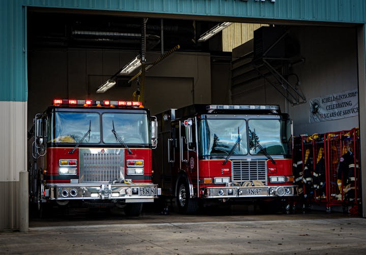 Two fire trucks parked in a fire station garage with a celebratory banner on the wall.