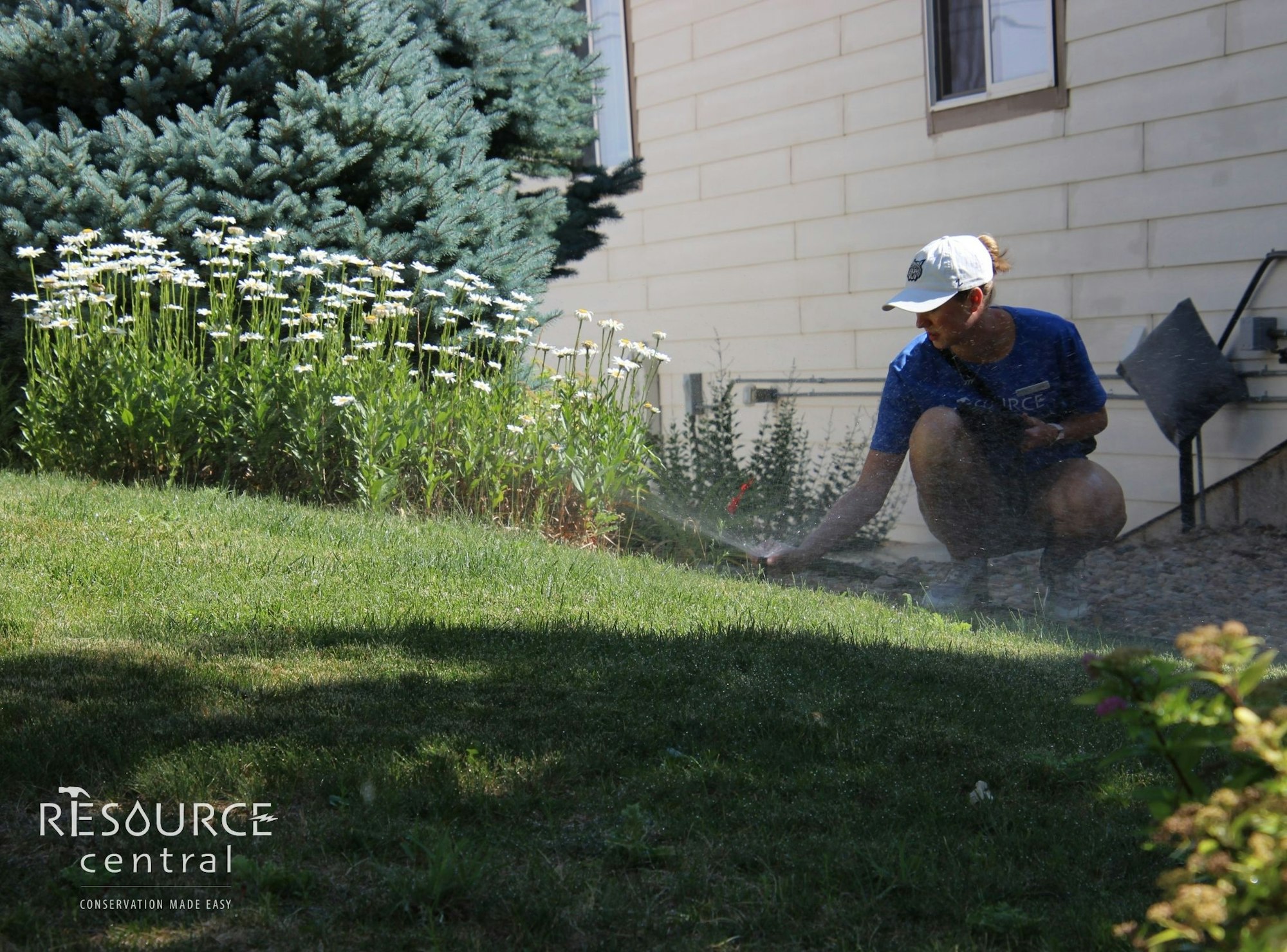 A person is watering plants and flowers in a garden beside a house.
