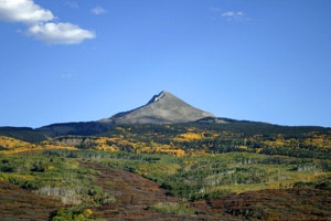 A mountain peak with a forested landscape showing autumn colors under a blue sky.