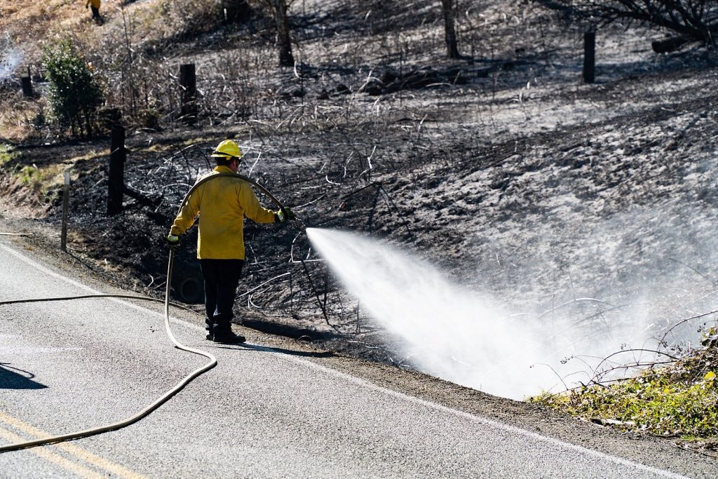 A firefighter dousing a charred area with water beside a road.
