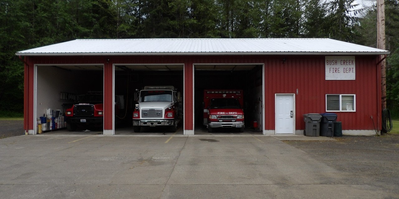 A red fire station with three open bays, two fire trucks visible, and a sign reading "BUSH CREEK FIRE DEPT."