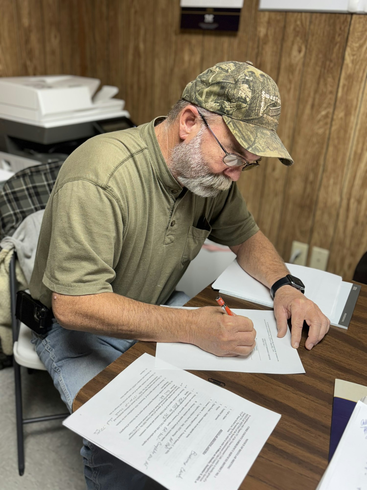 A man with a camouflaged cap is sitting at a table, writing on a document surrounded by papers.