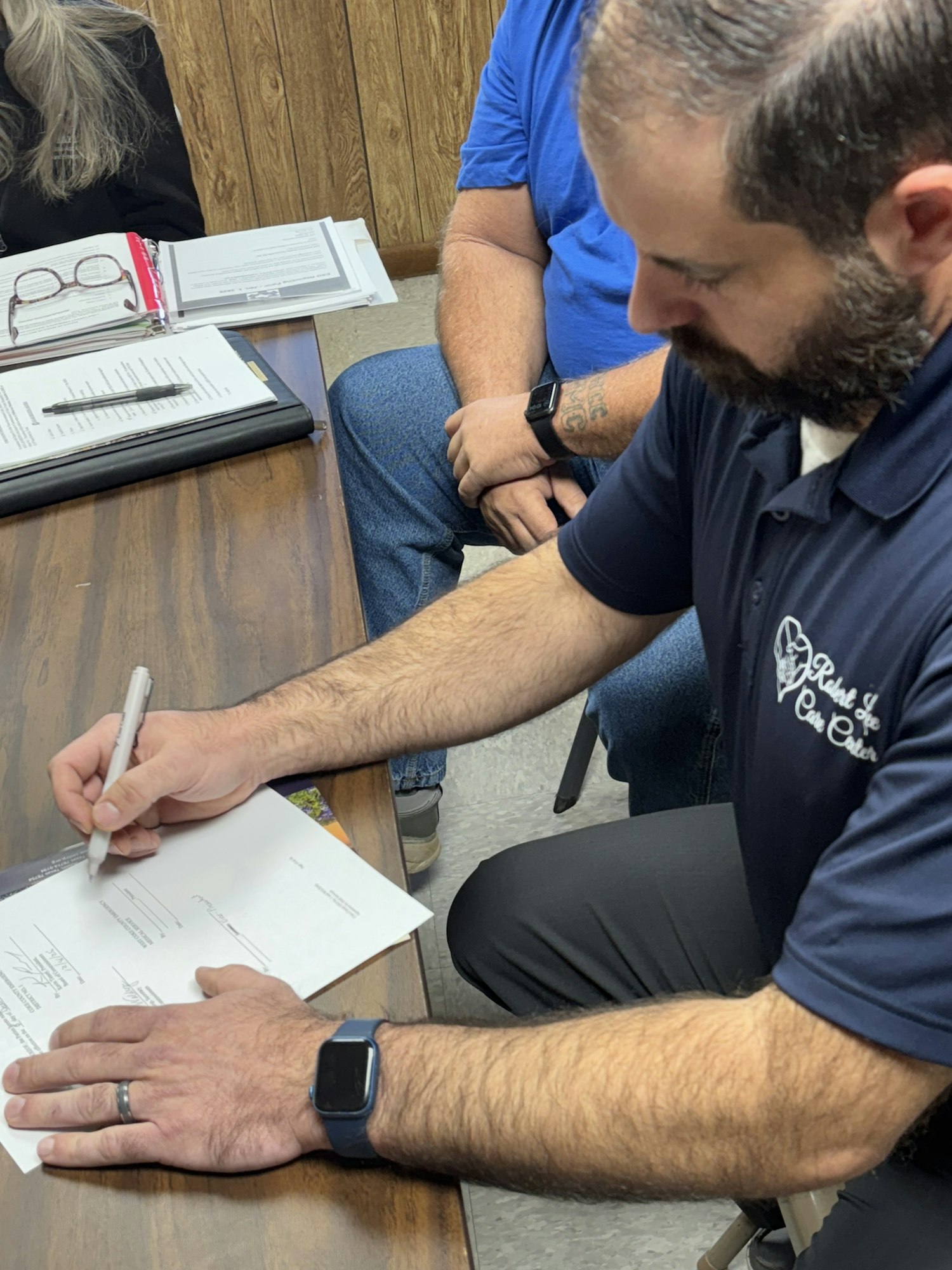 A person is signing a document at a table, with others observing in a casual setting. Multiple papers are visible.