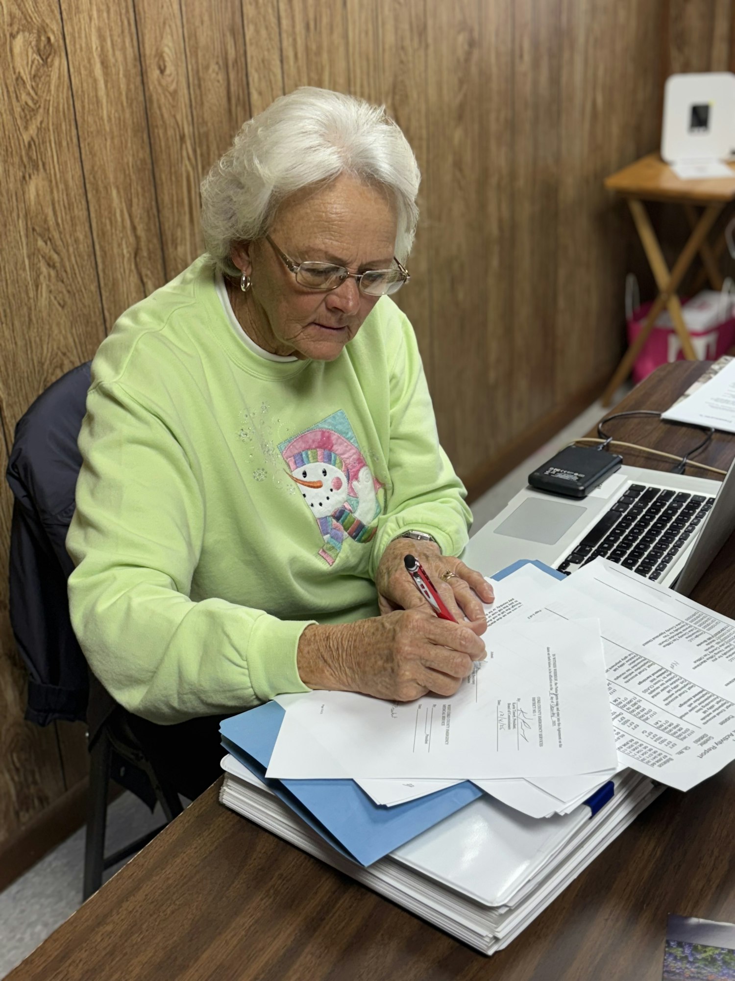 An older woman in a light green sweater is sitting at a desk, signing documents with papers and a laptop around her.