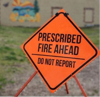 A bright orange sign reads: "PRESCRIBED FIRE AHEAD DO NOT REPORT." It indicates a controlled burn is in progress.