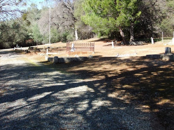 Rural outdoor scene with dirt ground, trees, shadows, and a fenced grave area.