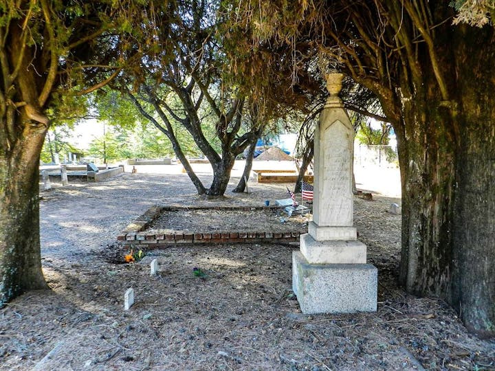 A sunlit cemetery with obelisk tombstone, small markers, trees, and fallen leaves.