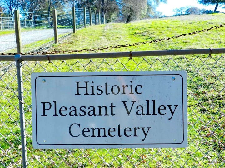 A sign reading "Historic Pleasant Valley Cemetery" attached to a chain-link fence with greenery in the background.