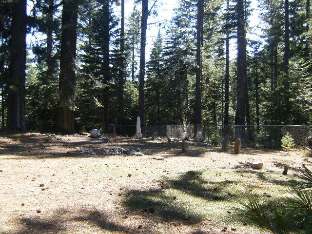 A fenced cemetery in a forest clearing with tall trees and scattered tombstones.