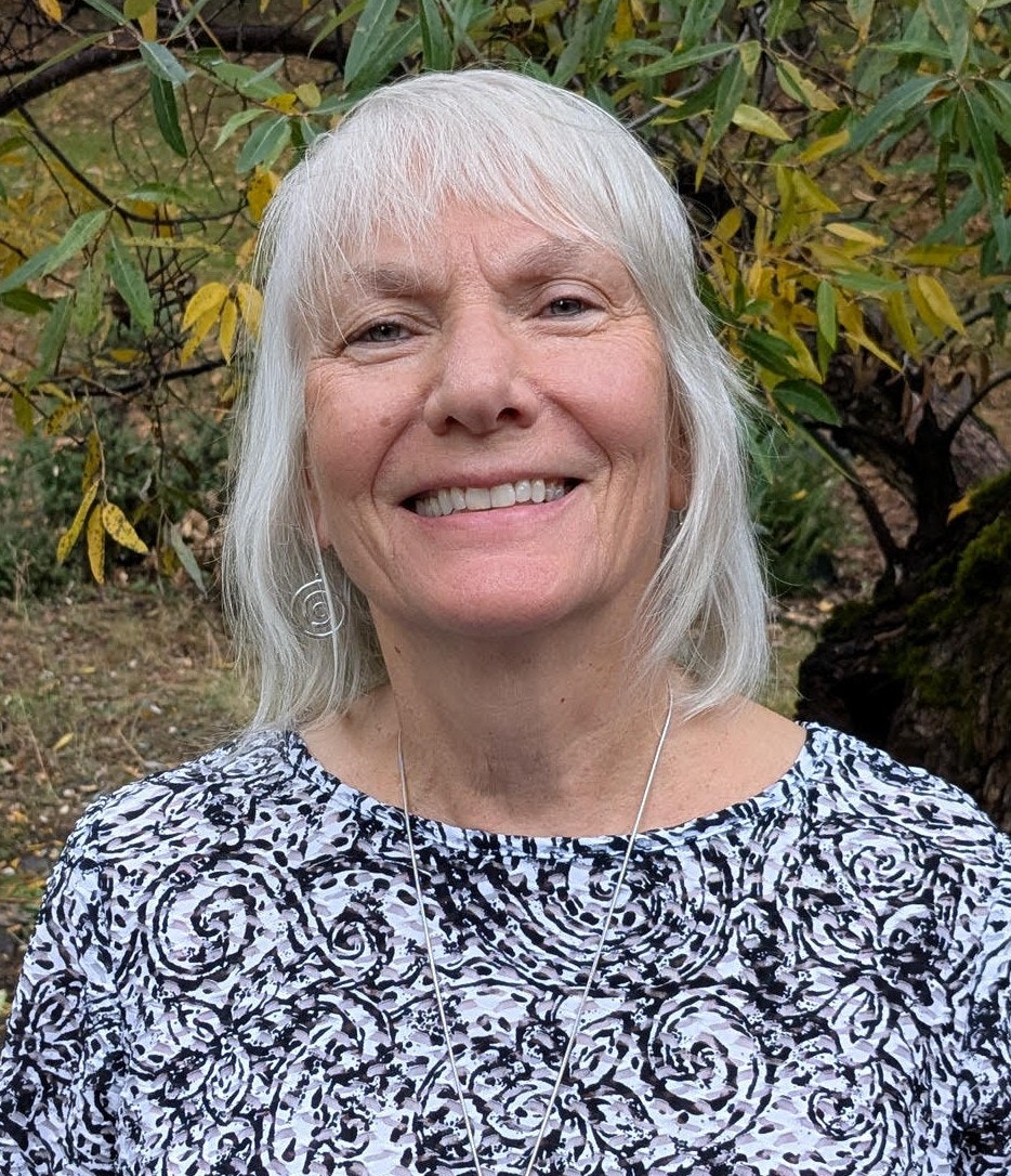 A smiling woman with gray hair is wearing a patterned top, standing outdoors with foliage in the background.