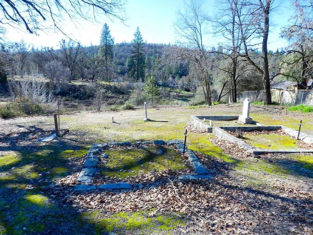 A tranquil, sunlit cemetery with sparse graves and trees, surrounded by a rural landscape.