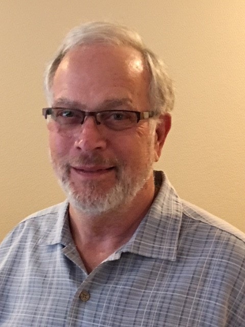 A man with glasses and a beard, wearing a checked shirt, smiles at the camera against a plain background.