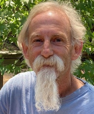 A person with a white beard and mustache, wearing a blue shirt, is outdoors with a leafy background.