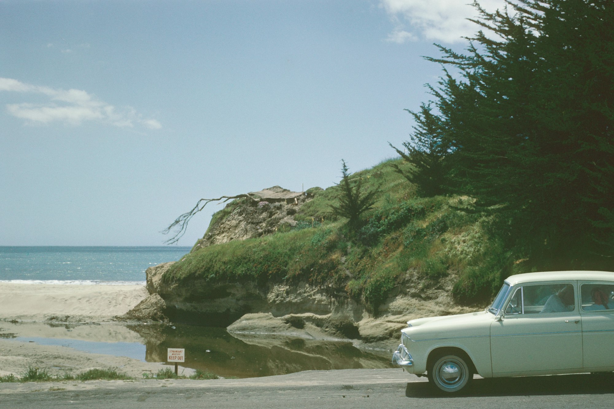 Vintage car parked by a coastal bluff with eroded structures and a "dangerous keep out" sign, under a clear blue sky.