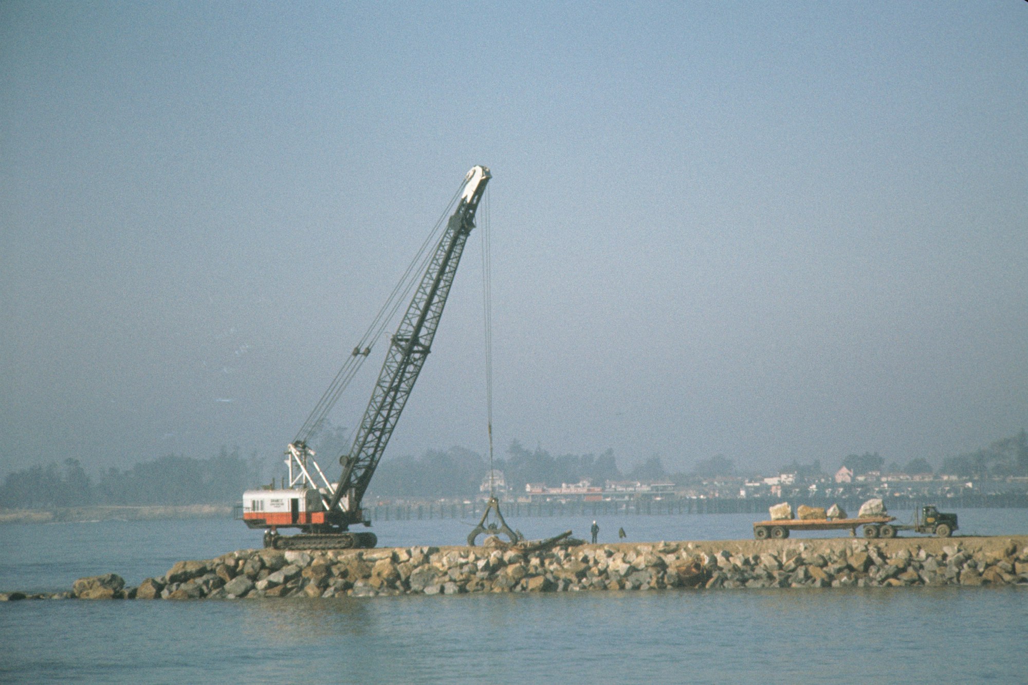 A crane on a breakwater lifting materials, with a water backdrop and some structures in the distance.