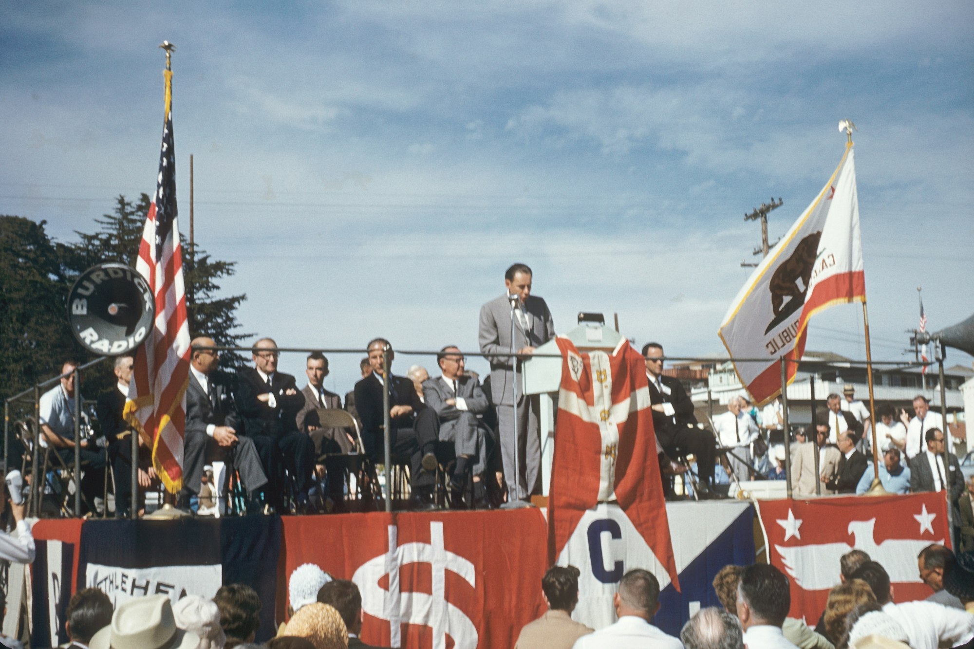 An outdoor event with a man speaking at a podium, audience, flags, and dignitaries seated on stage.