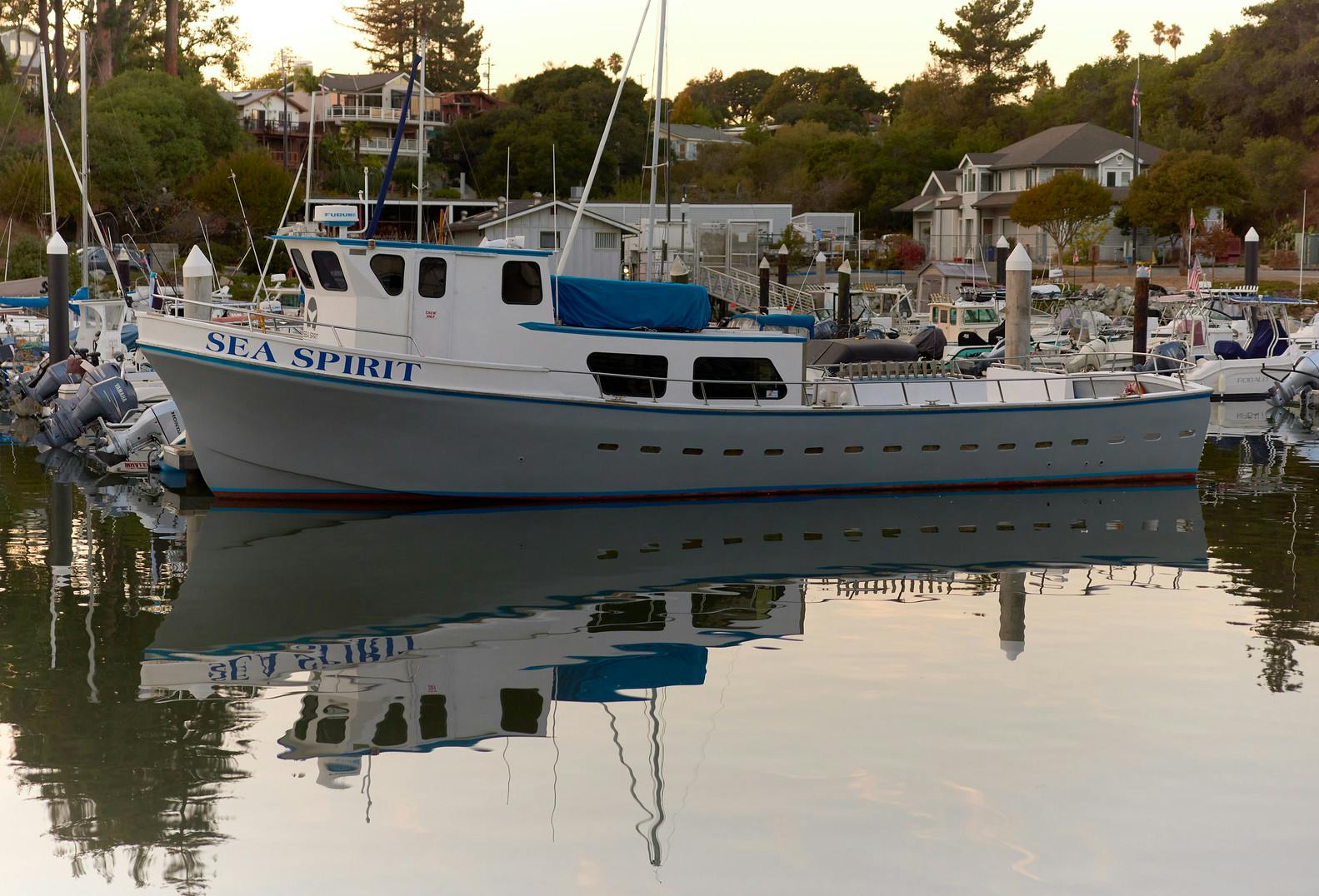 A blue and white boat named "SEA SPIRIT" docked in a calm marina with reflections on the water at sunset.