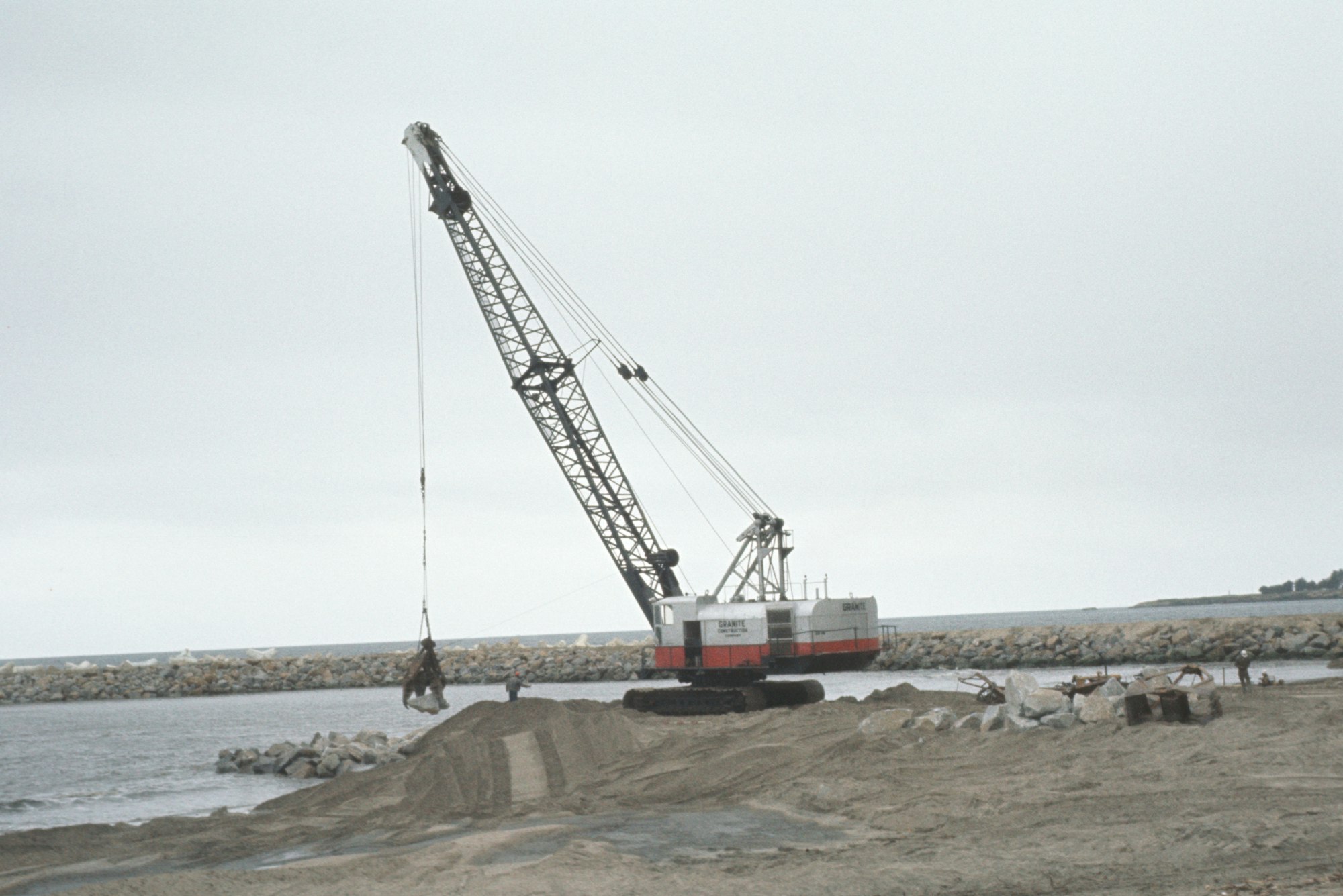 A crane on a sandy area near water, lifting a load, with a rocky breakwater in the background.