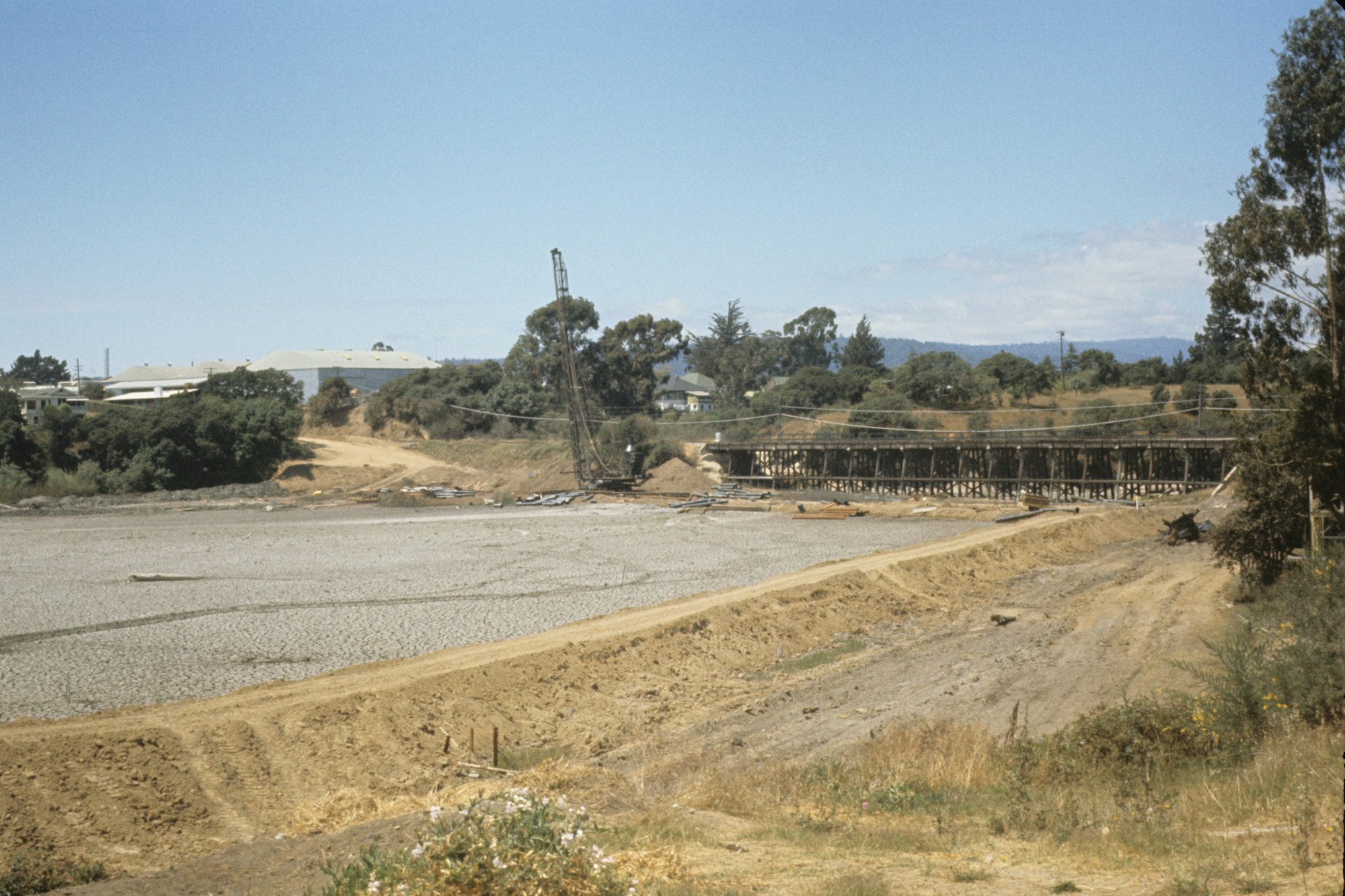 A dry riverbed with construction equipment, a partial bridge structure, and buildings surrounded by trees in the background.