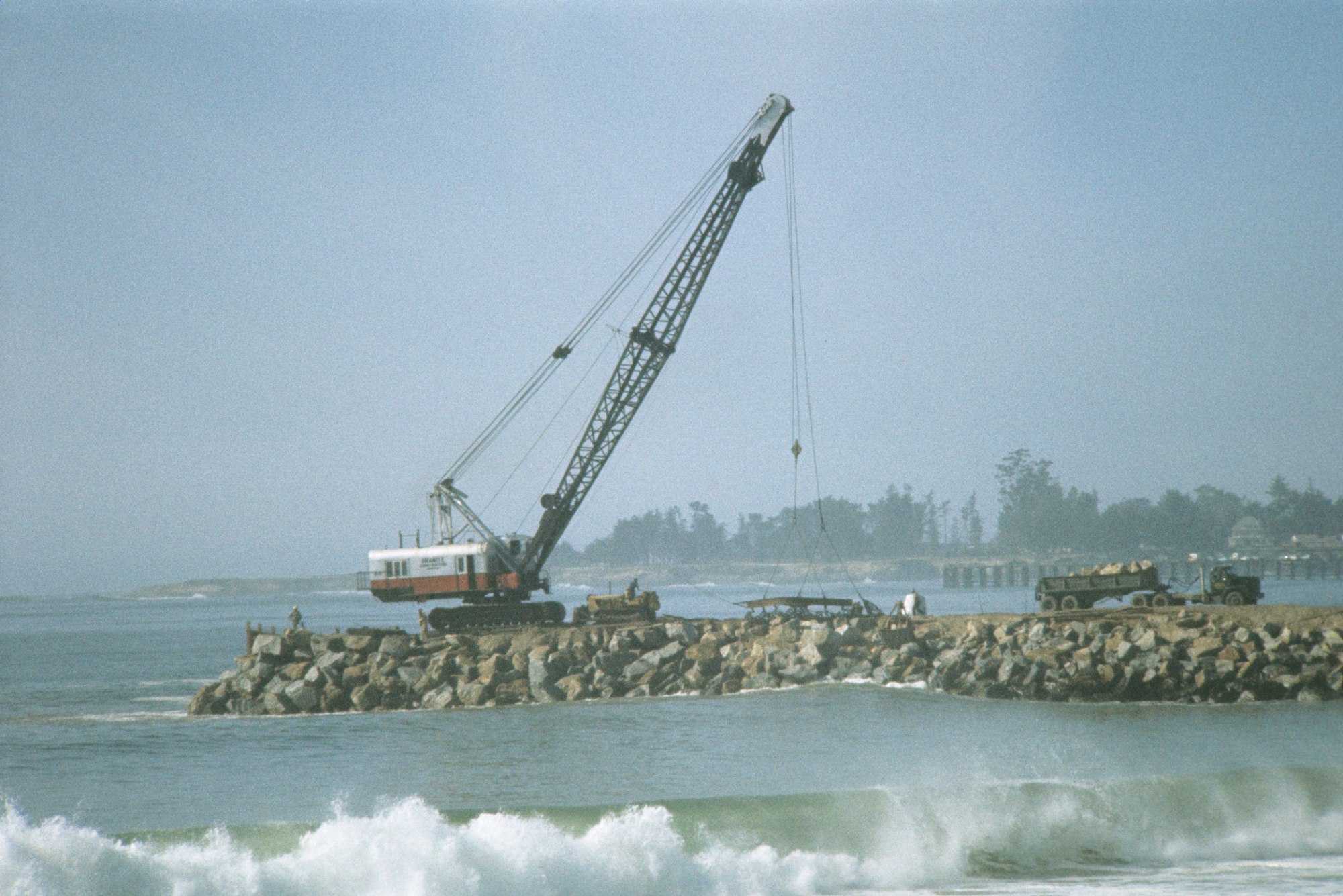 A crane on a rocky pier with a truck nearby, waves in the foreground, and a hazy sky.