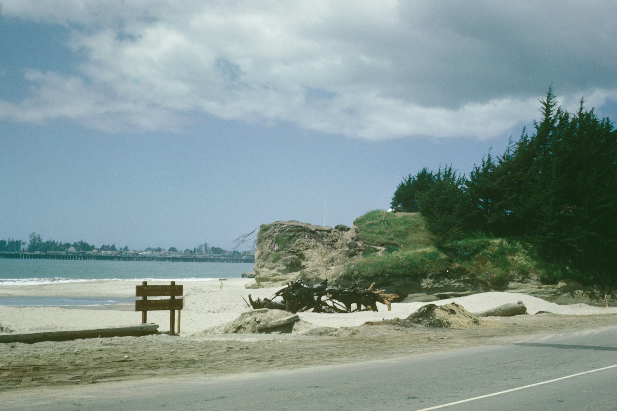 Sandy beach with a wooden sign, road in foreground, pier in the distance, and a cloudy sky.