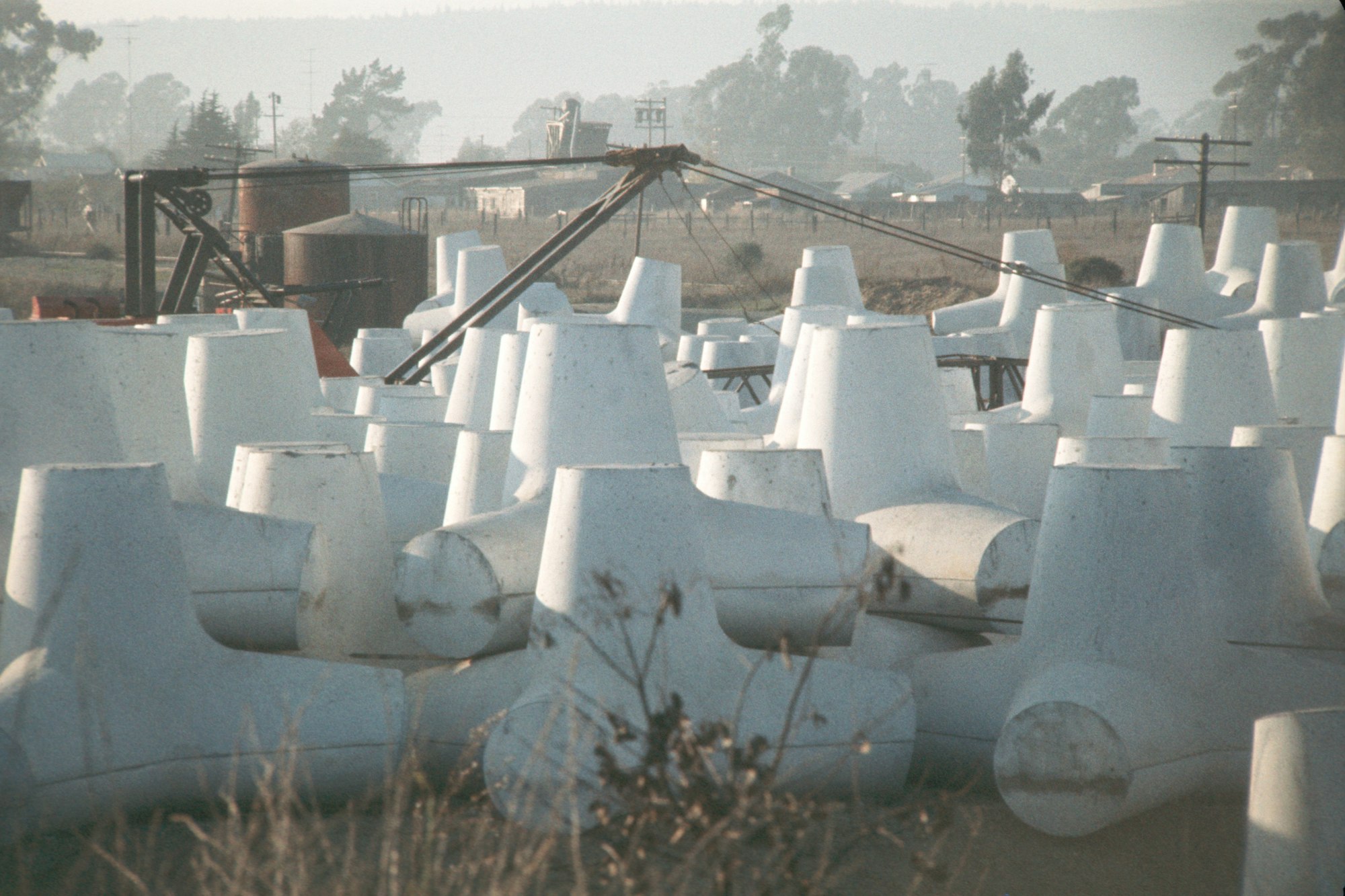 A field of large concrete pipes or culverts with industrial equipment in the background.