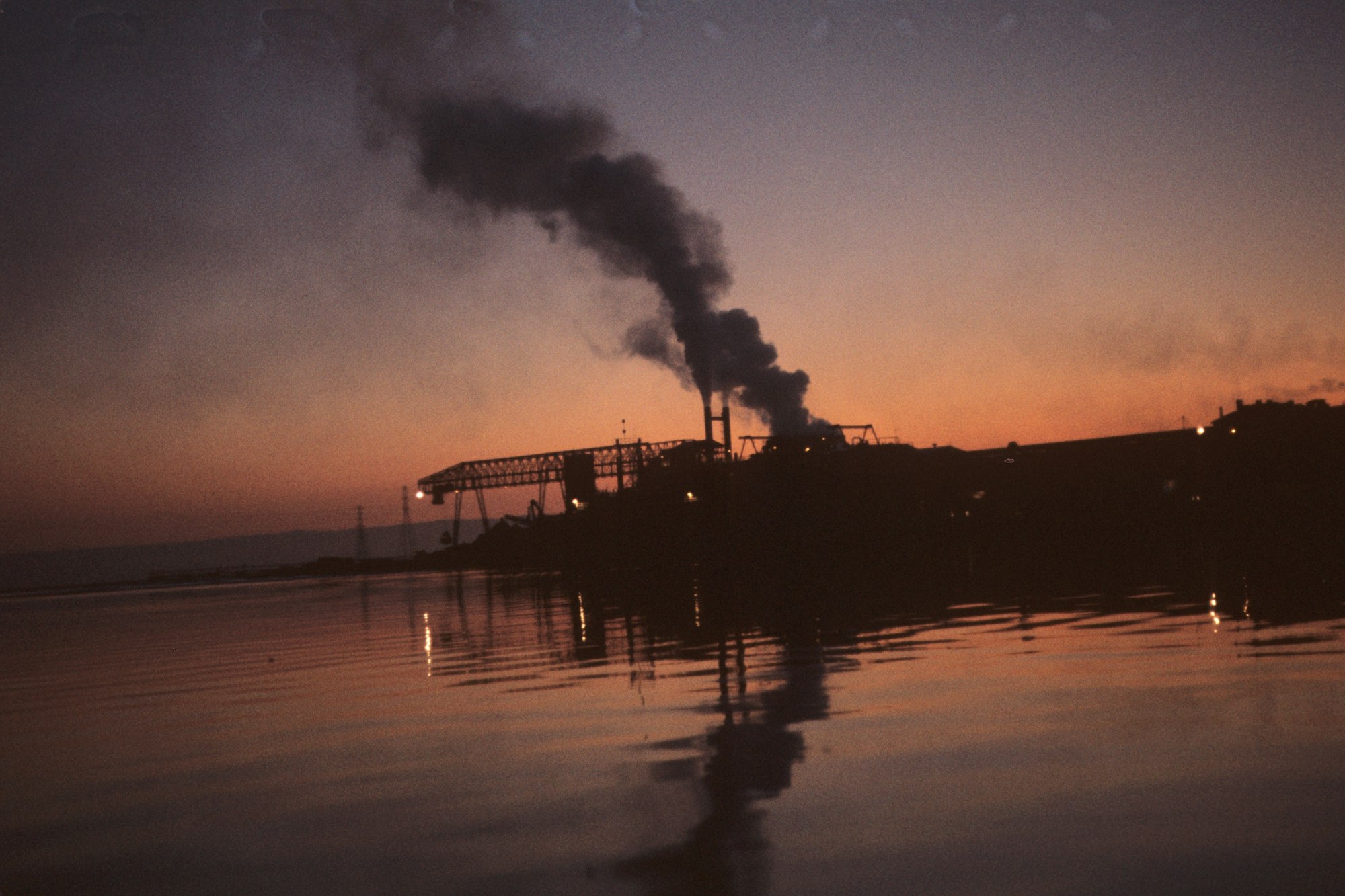 Industrial facility at dusk with smokestacks emitting smoke, reflected on water's surface.