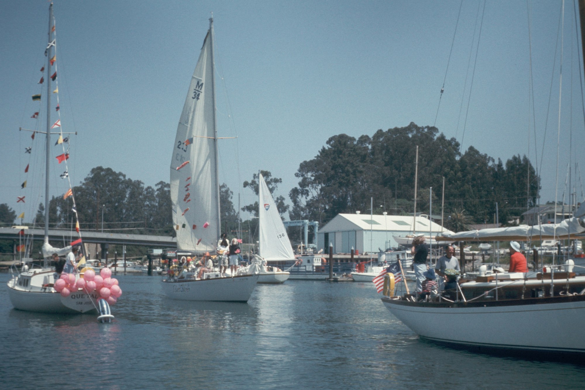 Sailboats with people on board, decorated with flags and balloons, floating in a marina under a clear sky.