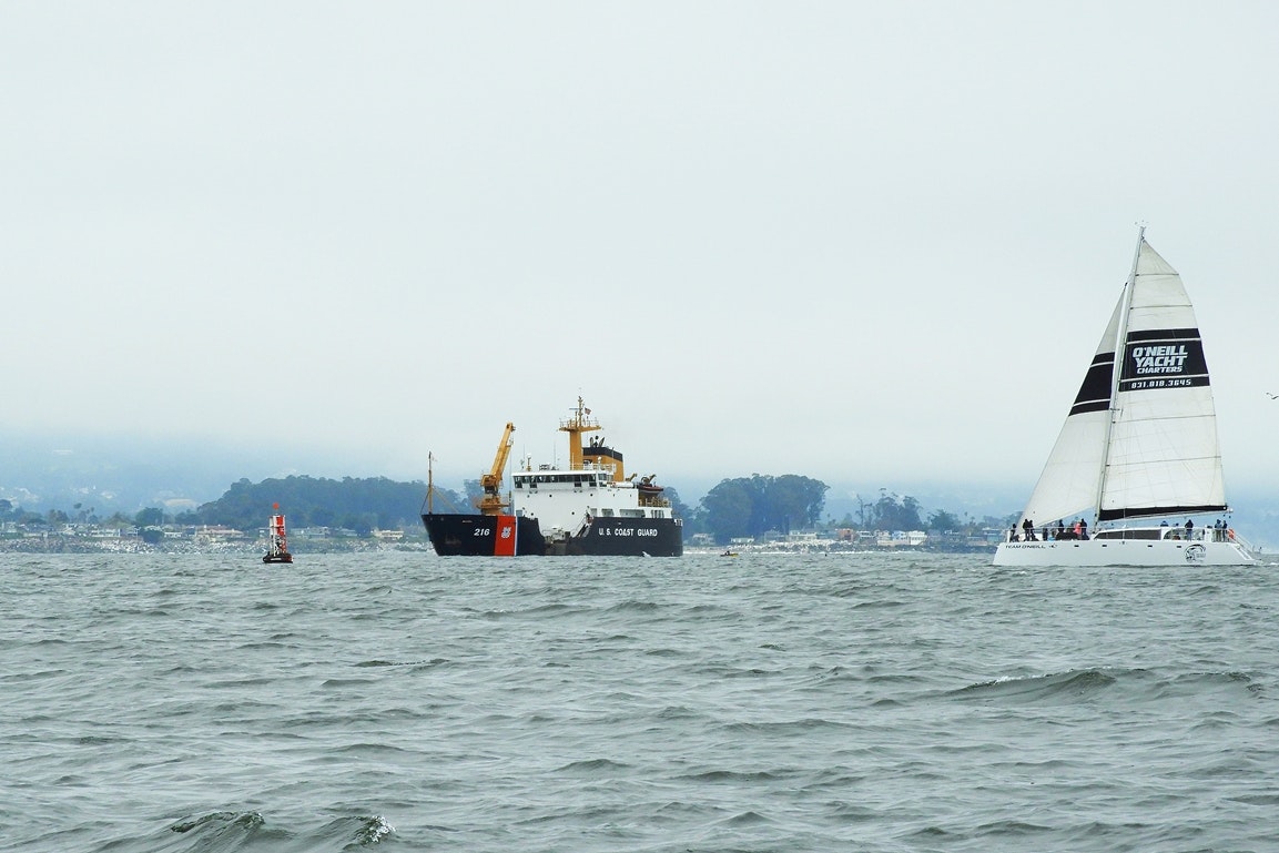 A Coast Guard ship and a sailboat on a hazy sea, with land in the distance.