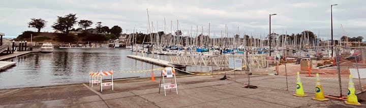 A marina with boats docked, surrounded by barriers and caution tapes, trees in the background, overcast sky.