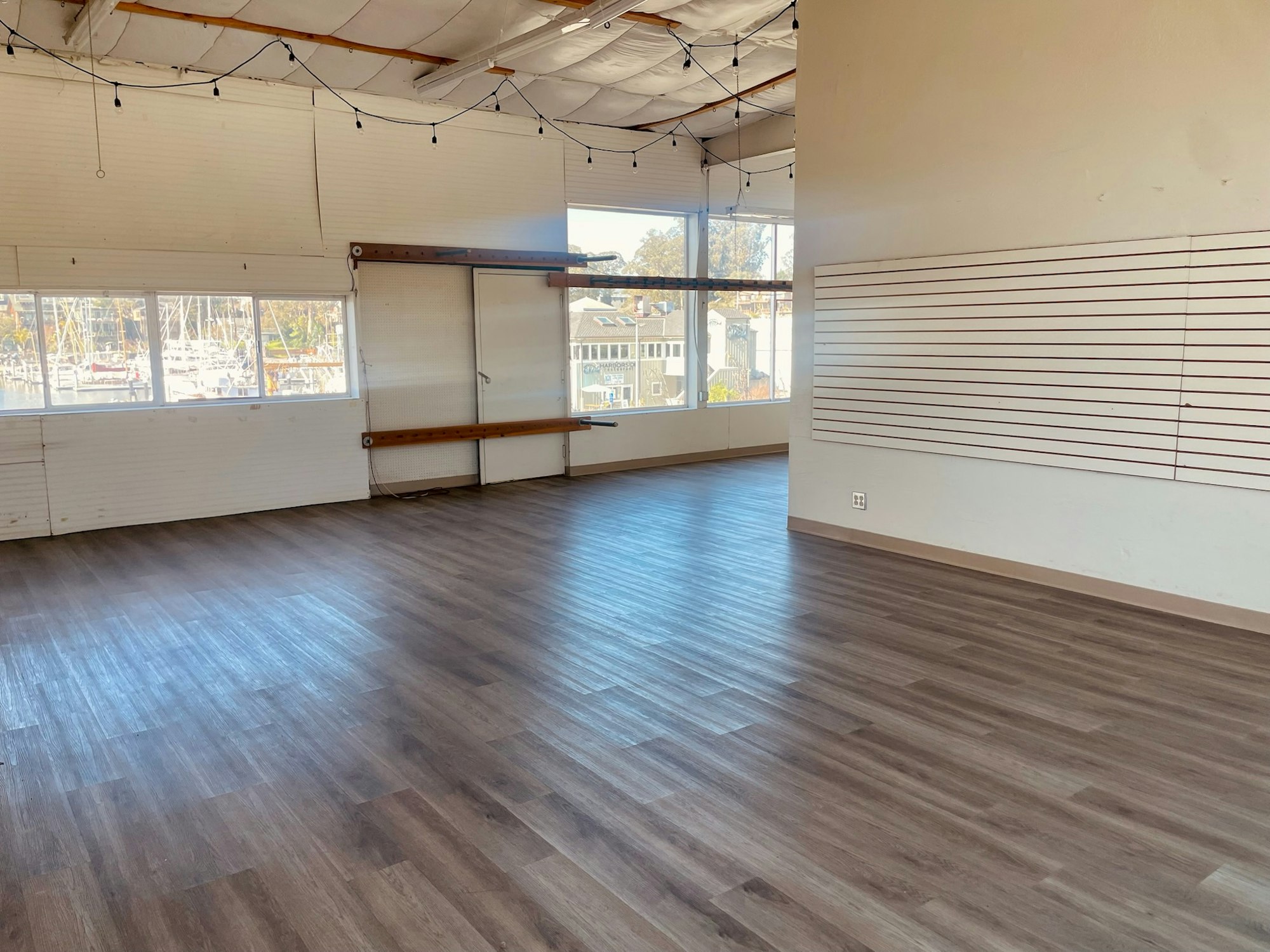 The image shows a spacious, bright room with wood-like flooring, windows overlooking a marina, and some shelves.