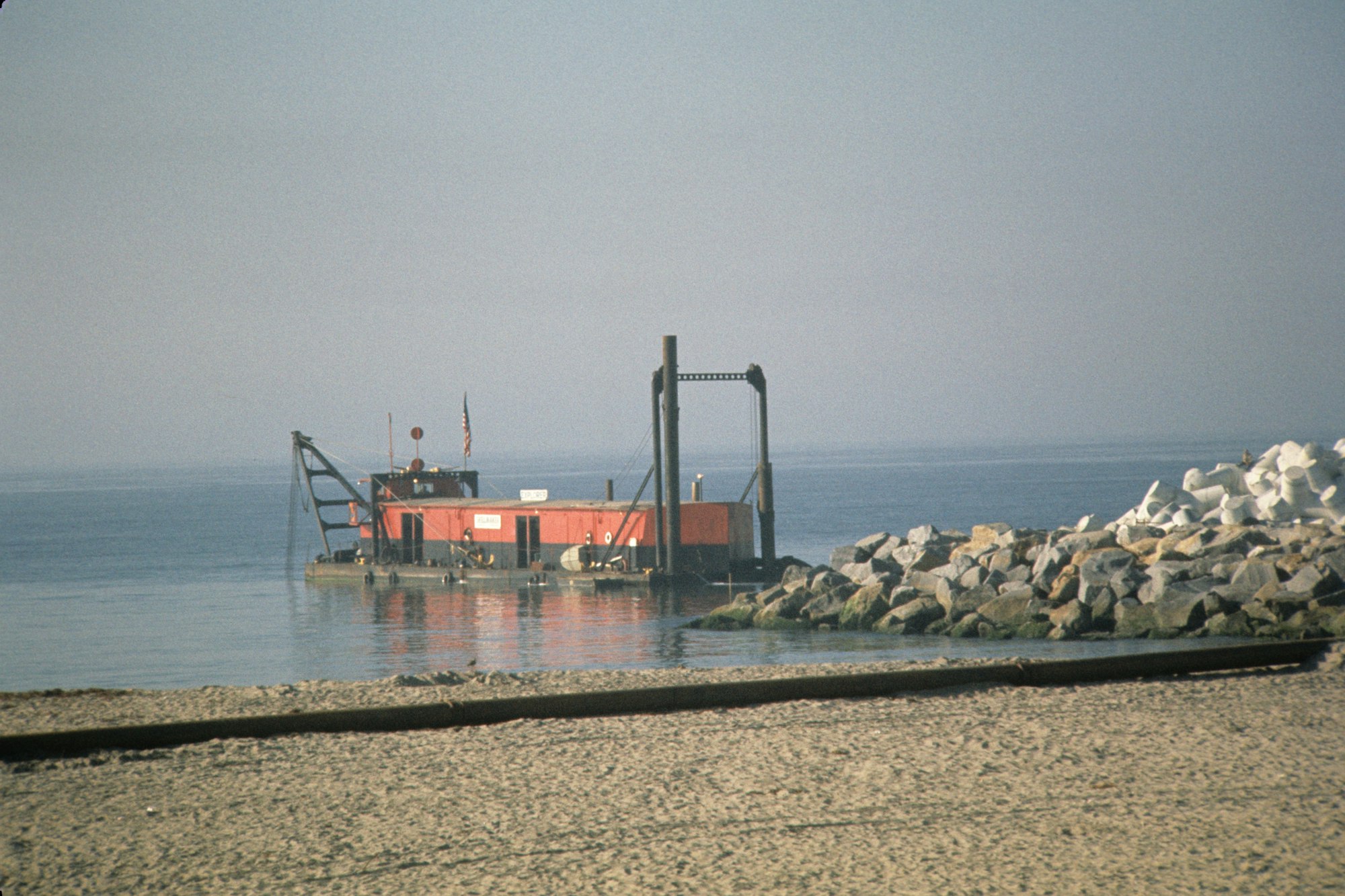 A barge with a crane at a calm seaside, next to a rocky jetty, with a sandy beach in the foreground.