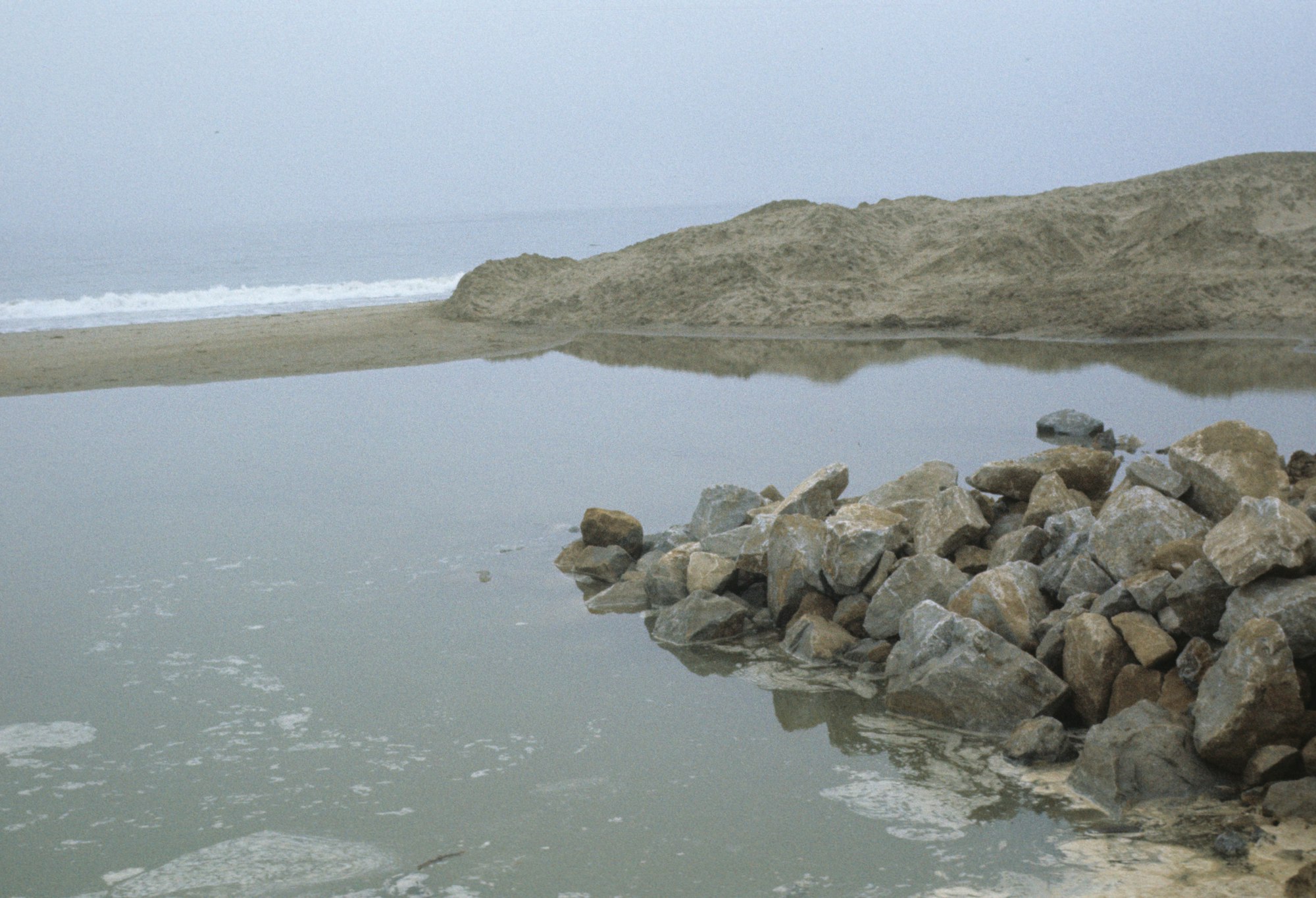 Coastal scene with rocks, sand dunes, water, and a misty ocean backdrop.