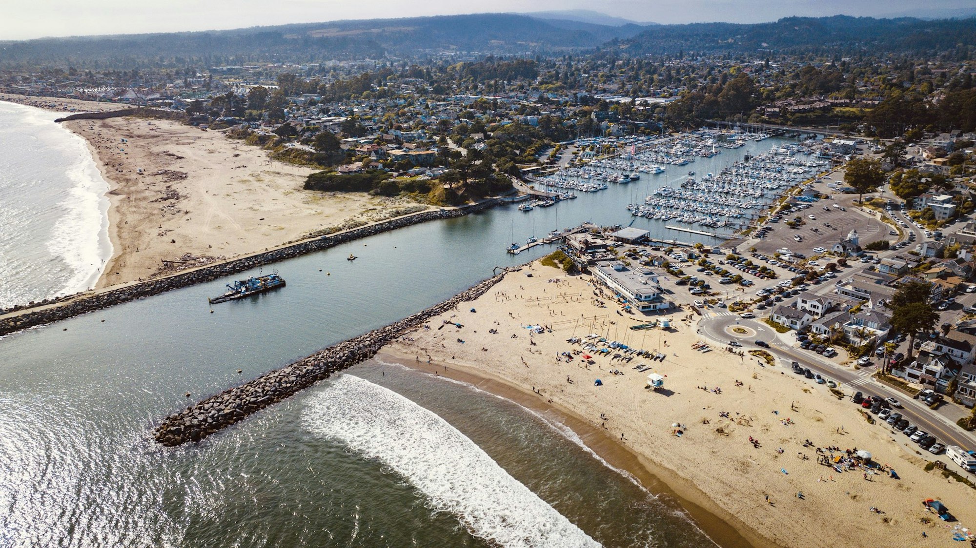 A scenic aerial view of a beach area with a marina, boats, and nearby residential areas, showcasing sand, waves, and people enjoying the beach.