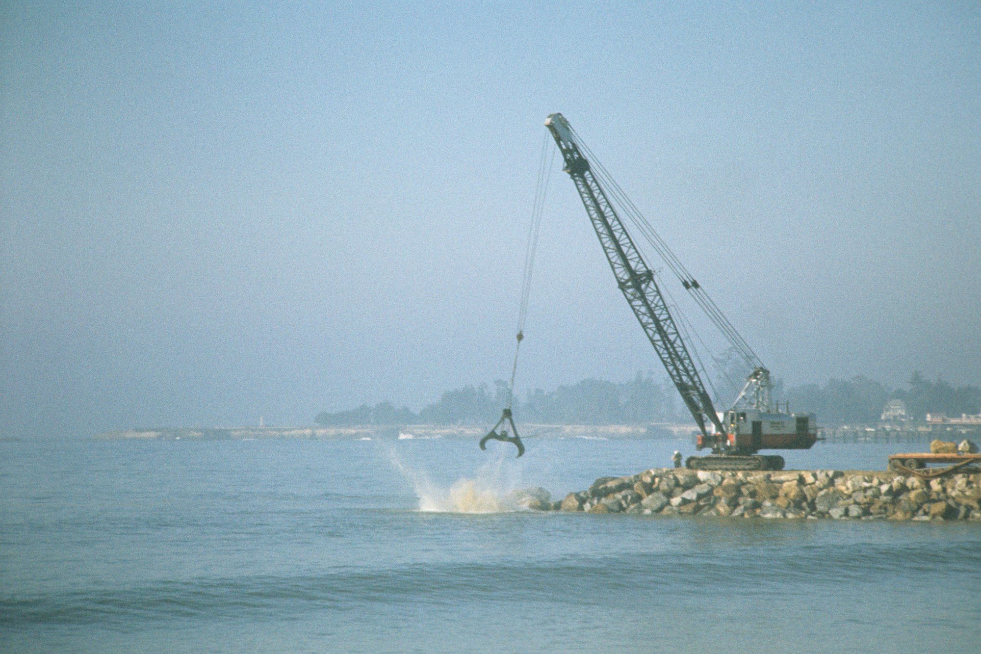 A crane on a breakwater lifting a large object, creating a splash in the water, with hazy background.