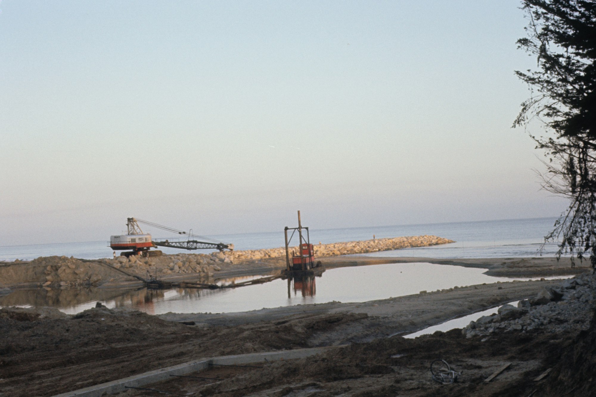 Construction equipment by the shore with a partially built jetty extending into calm waters, under a clear sky.