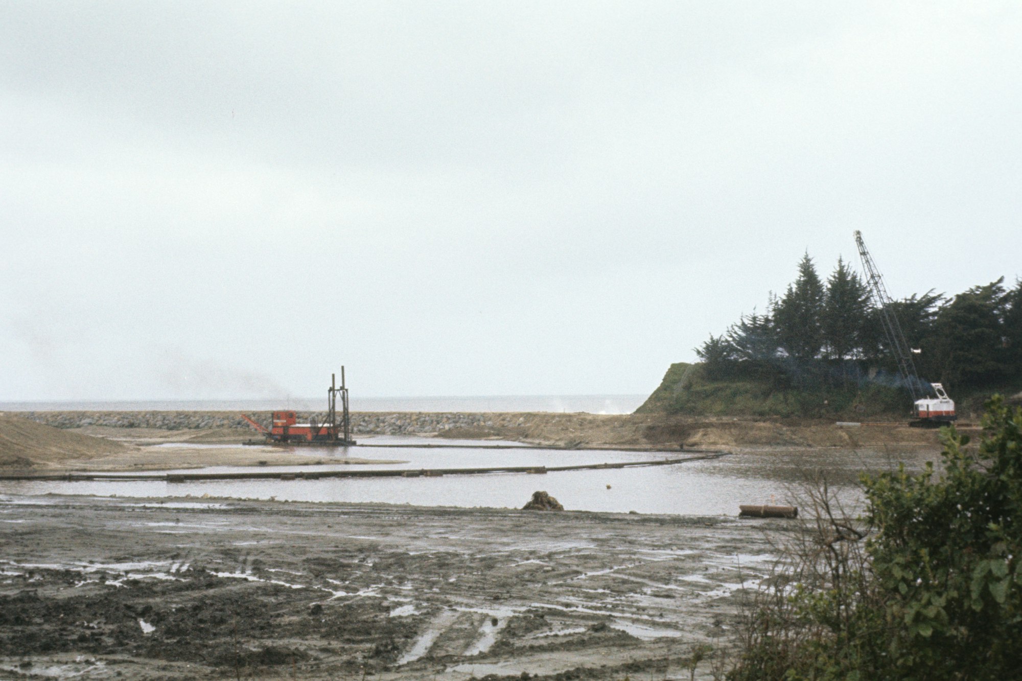 Coastal construction site with machinery and a water body, under a cloudy sky.