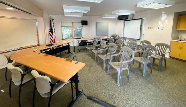 A conference room with tables, chairs, an American flag, and a projector screen.