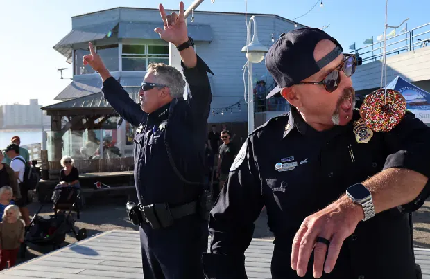 Two police officers perform on stage, one dancing and the other holding a donut, entertaining a crowd by the waterfront.