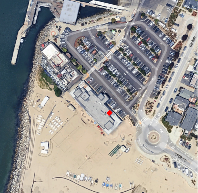 An aerial view showing a beach area with a pier, parking lots, buildings, and a roundabout near the shoreline.