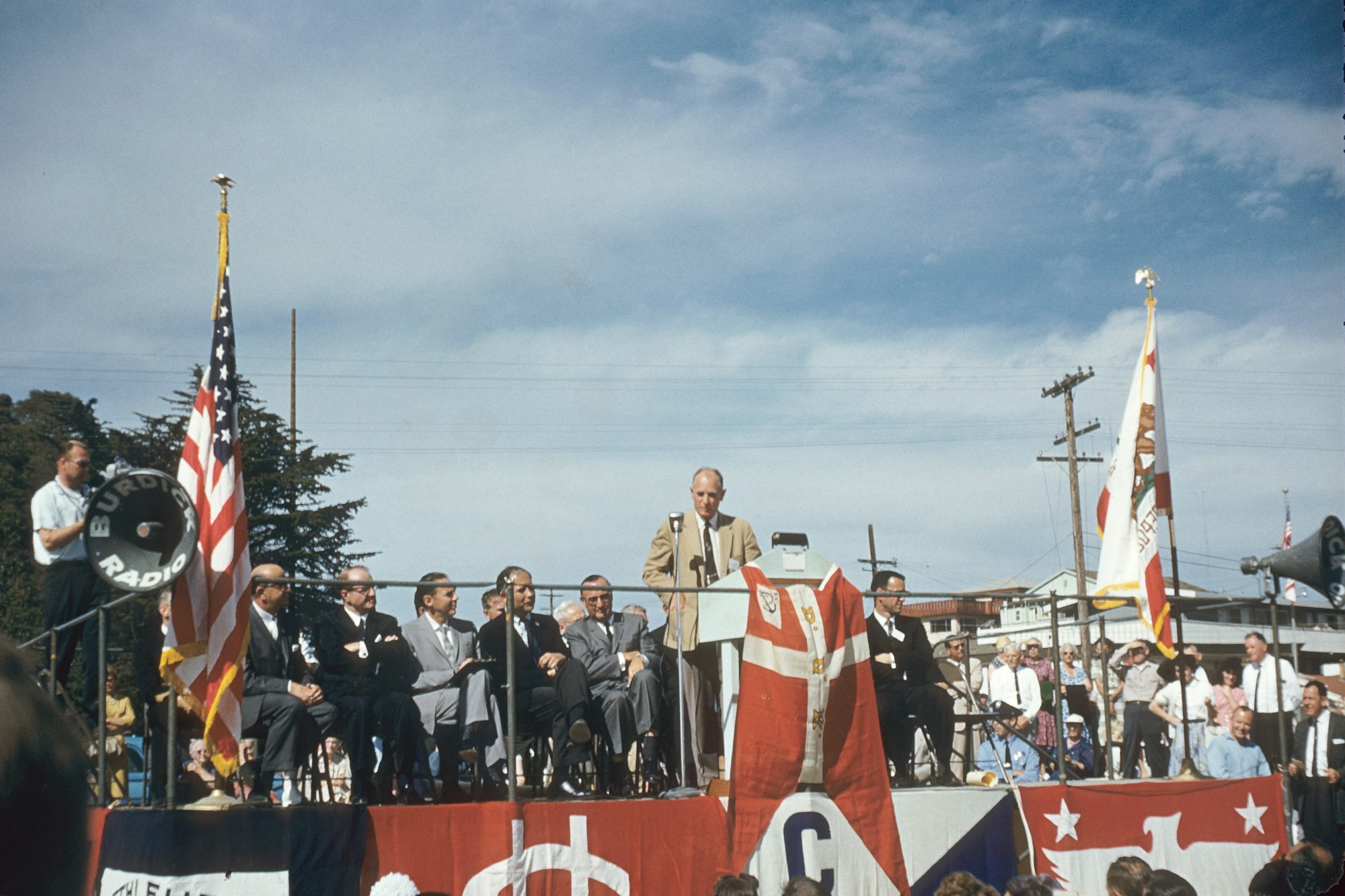 A man speaks at a podium during a ceremony with seated officials, U.S. flags, and a "Burdick Radio" microphone in the foreground.